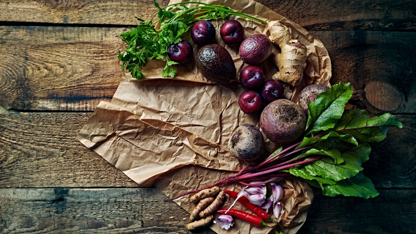 Verschiedene Gemüsesorten, darunter Rüben, violettes Gemüse, Ingwer, Chilischoten und Petersilie, werden auf zerknülltem braunem Papier auf einer Holzoberfläche präsentiert – perfekt für Anfänger, die vegane Rezepte ausprobieren möchten.